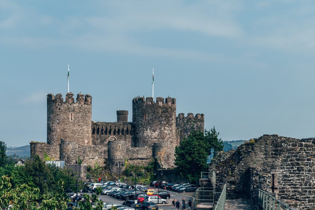 Stone towers of Conwy Castle in Wales above a car park and walls under a clear sky.