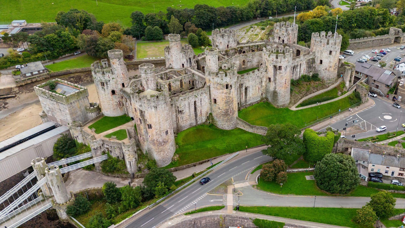 Drone view of Conwy Castle and suspension bridge in Conwy, Wales, with rugged towers and green ramparts.