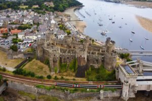 Aerial view of Conwy Castle in Wales beside the estuary, village rooftops, marina, bridge, and passing train.