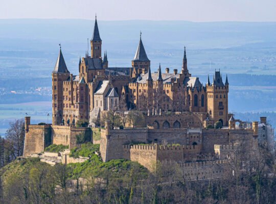 Hohenzollern Castle stands majestically atop a wooded hill in Germany, surrounded by lush scenery.