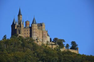 Hohenzollern Castle rises above forested hills in Germany under a clear blue sky.