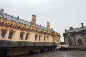 Stirling Castle courtyard on a cloudy, rainy day, showing historic yellow stone architecture and people below.
