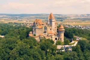 Aerial of Kreuzenstein Castle amid forested hill and patchwork fields near Leobendorf, Lower Austria, under warm summer light.