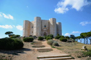 Octagonal limestone Castel del Monte in Apulia, Italy, viewed from steps under blue sky and Mediterranean scrub.