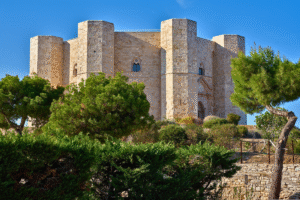 Castel del Monte in Puglia, Italy, octagonal medieval fortress rising above pines under a clear blue sky.
