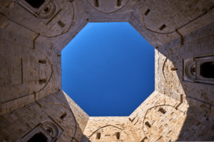 View up through octagonal courtyard of Castel del Monte, Apulia, revealing stone walls and deep blue sky.