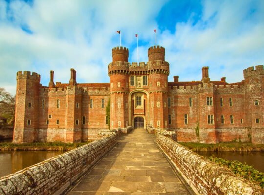 Stone bridge to Herstmonceux Castle, a moated red-brick fortress under bright skies in East Sussex, England.