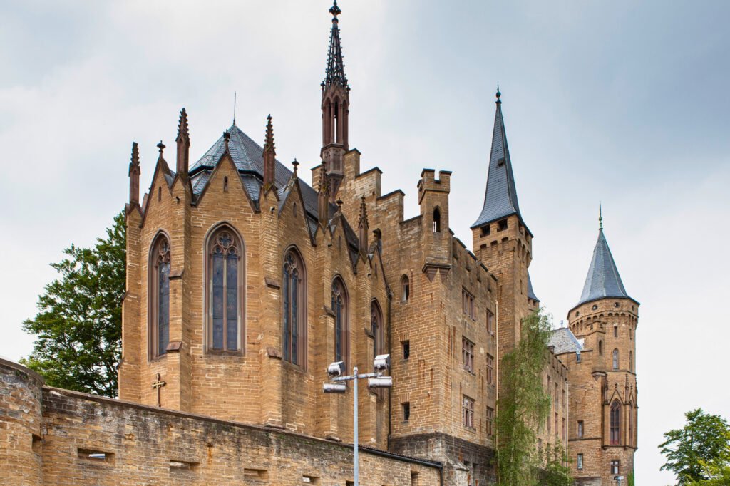 Sandstone Hohenzollern Castle with Gothic spires and turrets rising over fortress walls under a cloudy sky.