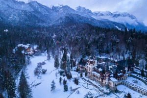 Aerial view of Peleș Castle in snowy Sinaia, Romania, framed by Carpathian Mountains and forest.