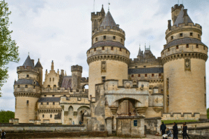 Front view of Pierrefonds Castle in Oise, France, with towering turrets and stone gate under cloudy sky.