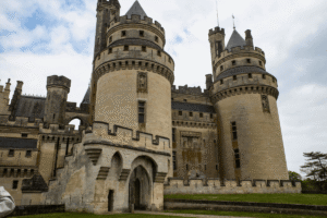 Stone towers and gate of Château de Pierrefonds in Oise, France, under cloudy sky.