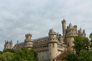 Château de Pierrefonds rising above trees under overcast sky, showcasing round towers and medieval revival architecture.