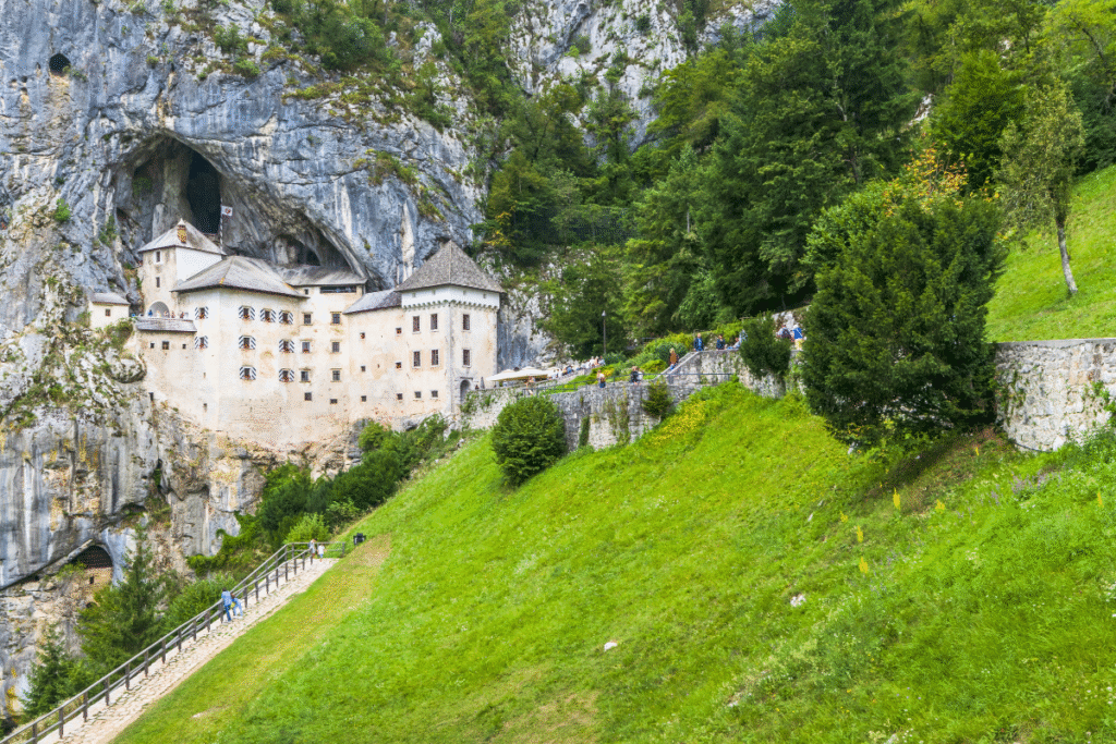 Predjama Castle in Slovenia built into a cliff, cave opening behind, visitors walking up the green hillside.