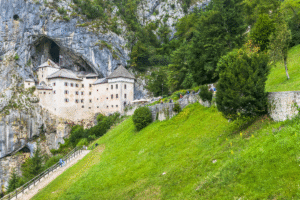 Predjama Castle in Slovenia built into a cliff, cave opening behind, visitors walking up the green hillside.