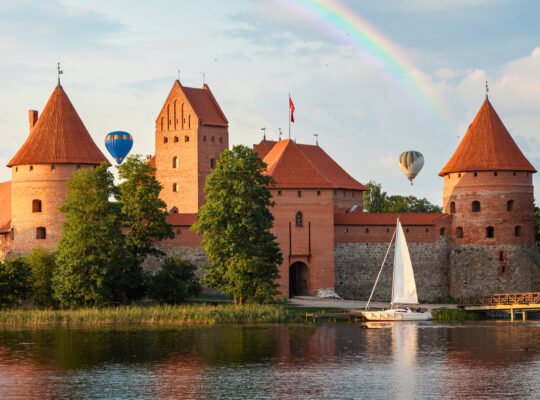 Trakai Island Castle at sunset with rainbow, hot air balloons, and sailboat on Lake Galvė.