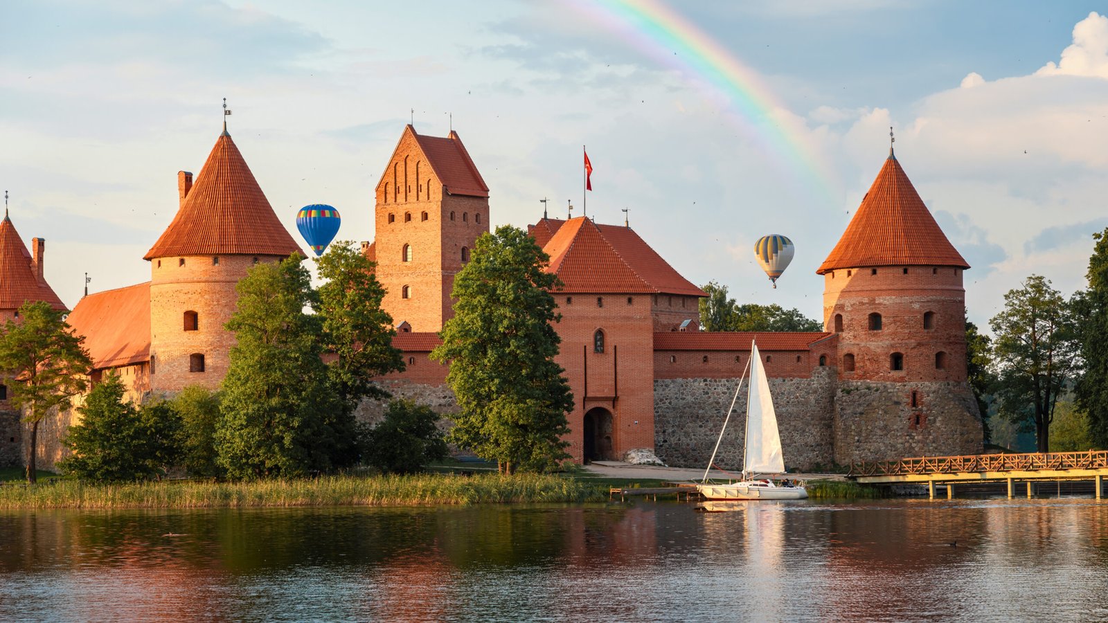 Trakai Island Castle at sunset with rainbow, hot air balloons, and sailboat on Lake Galvė.