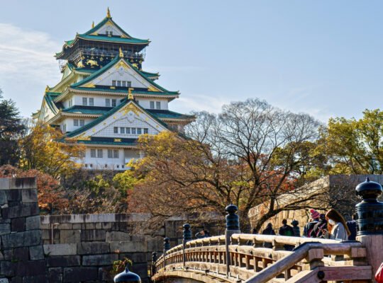 Osaka Castle framed by autumn trees and stone bridge, visitors crossing