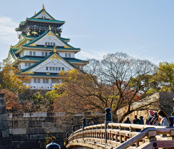Osaka Castle framed by autumn trees and stone bridge, visitors crossing
