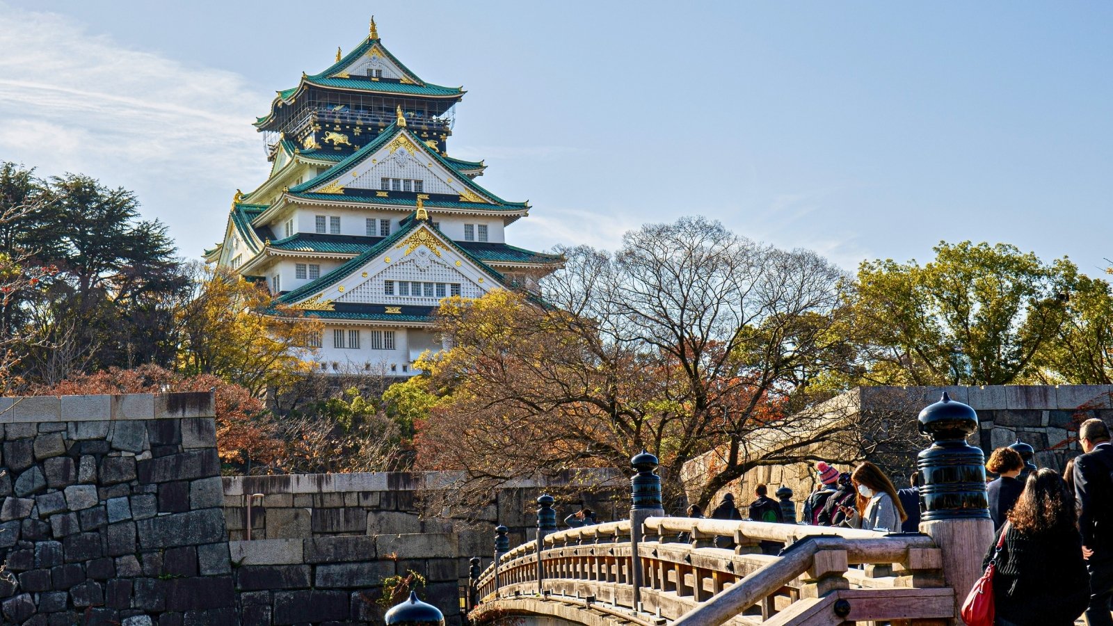 Osaka Castle framed by autumn trees and stone bridge, visitors crossing