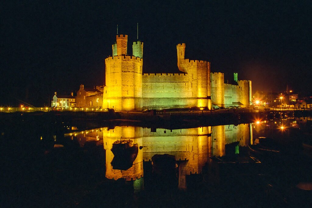 Illuminated Caernarfon Castle reflecting in calm harbor water at night