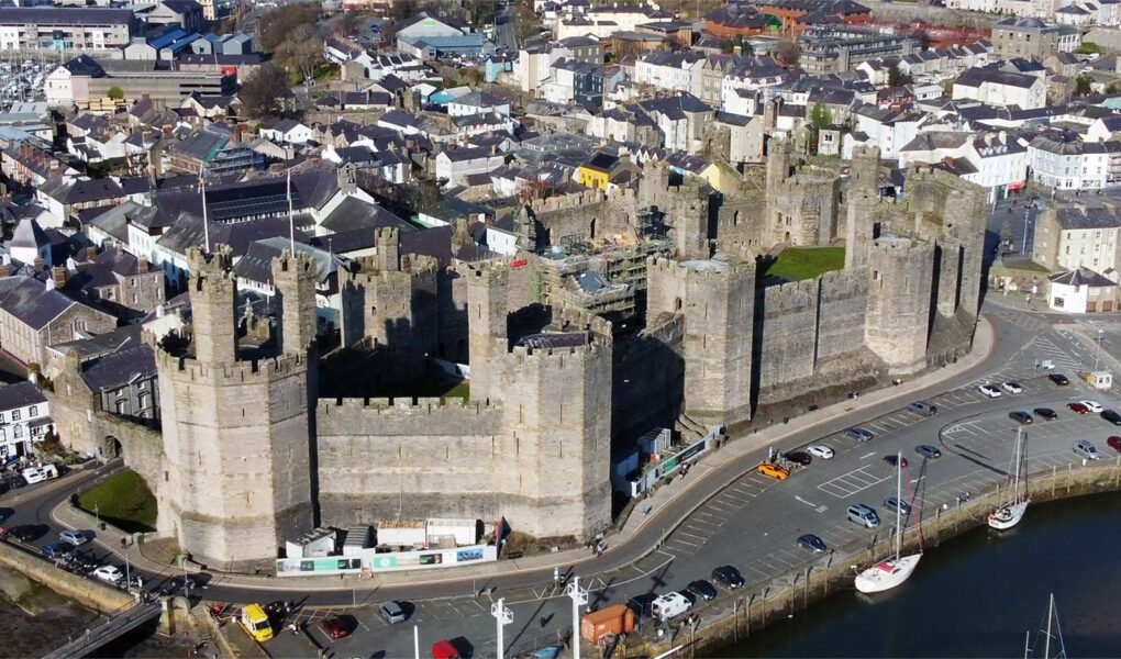 Aerial view of Caernarfon Castle with town and harbour in sunlight.