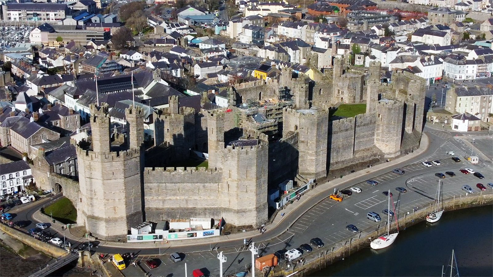 Aerial view of Caernarfon Castle with town and harbour in sunlight.