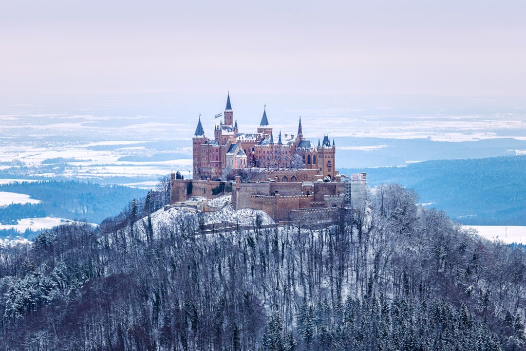 Snow-covered Hohenzollern Castle perched on forested hill overlooking winter plains