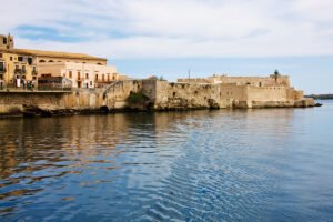 Maniace Castle along sunlit seaside walls, calm water reflection