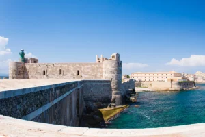 Maniace Castle on rocky Sicilian coastline under bright blue sky