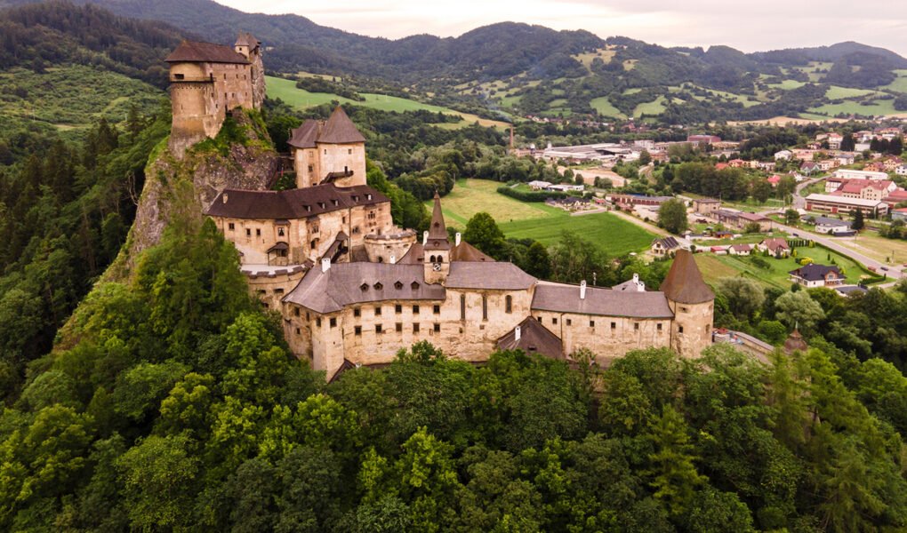 Aerial view of Orava Castle perched on rocky hill above forest and village