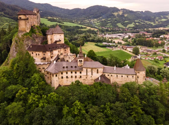 Aerial view of Orava Castle perched on rocky hill above forest and village
