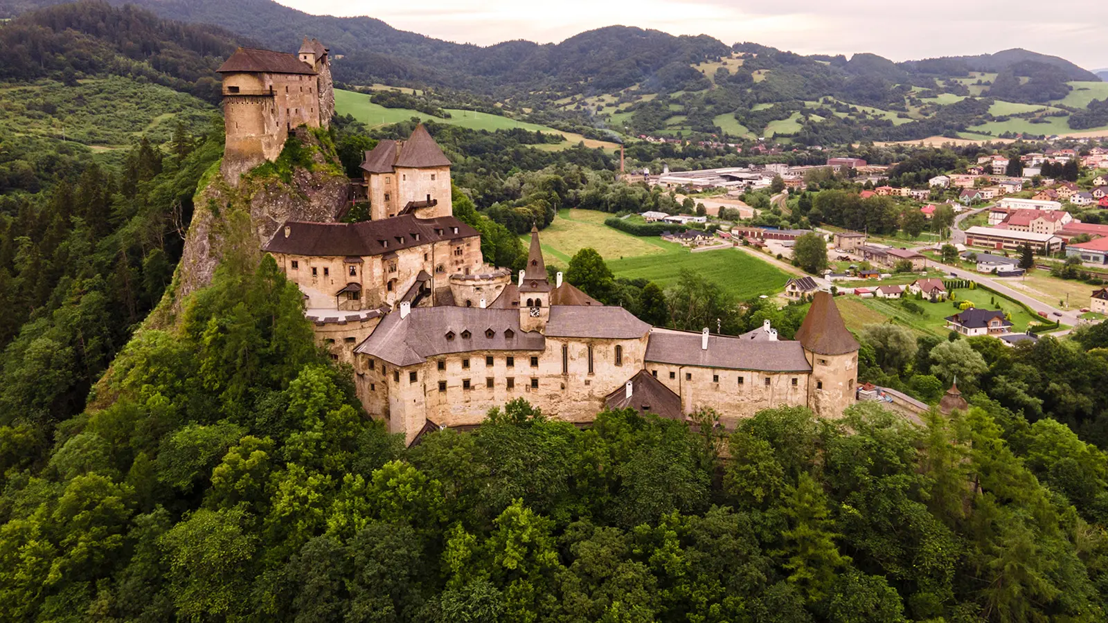 Aerial view of Orava Castle perched on rocky hill above forest and village