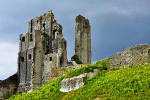 Ruined stone towers of Corfe Castle on green hillside under stormy sky