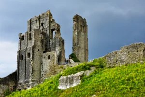 Ruined stone towers of Corfe Castle on green hillside under stormy sky