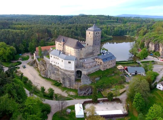 Aerial view of Kost Castle perched on sandstone cliff beside a reflective pond and forest