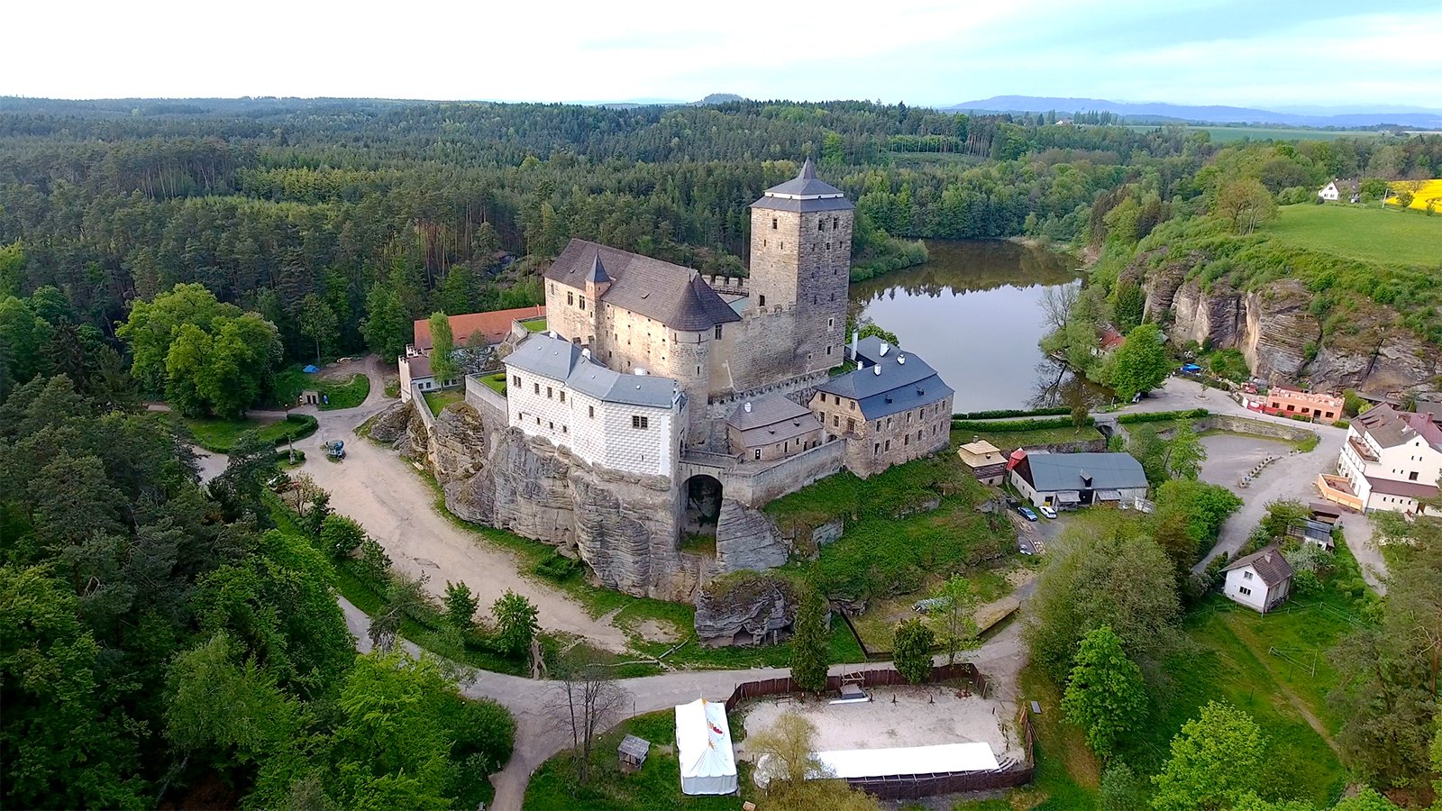 Aerial view of Kost Castle perched on sandstone cliff beside a reflective pond and forest