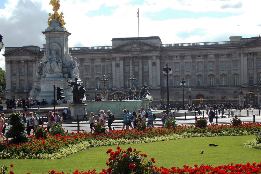 Buckingham Palace facade behind Victoria Memorial and red flowerbeds, visitors strolling