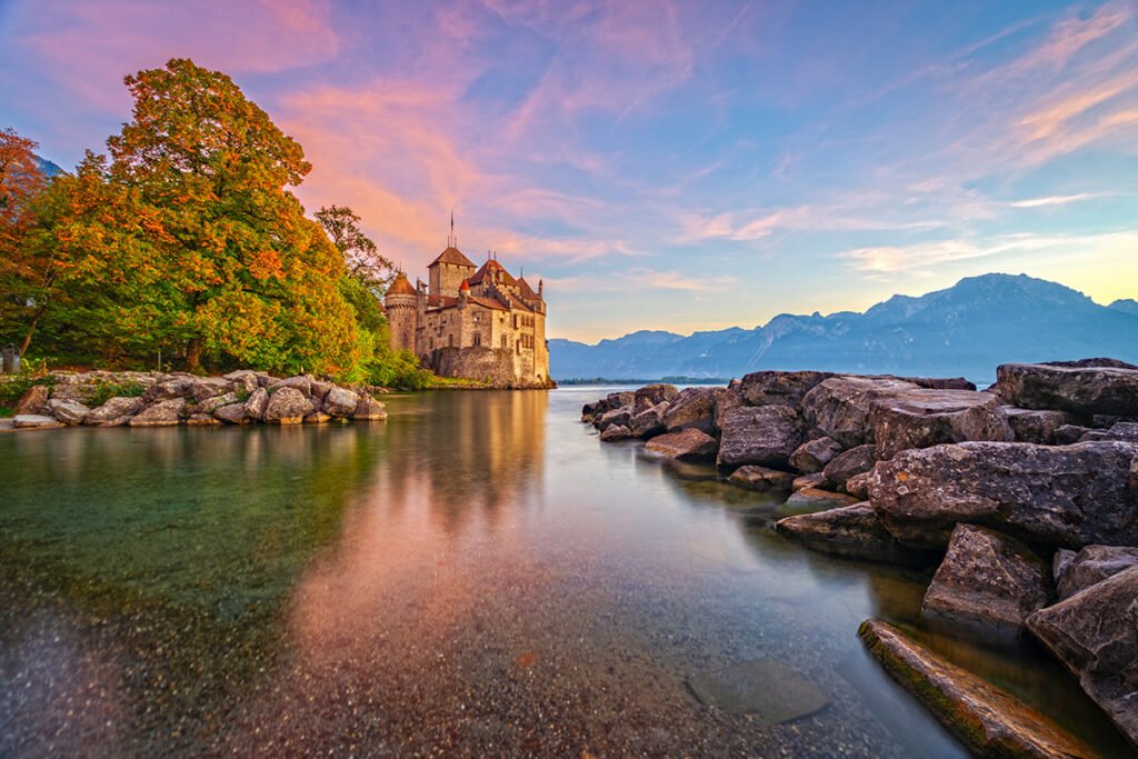 Chillon Castle beside Lake Geneva at colorful sunset, autumn trees and Alpine backdrop