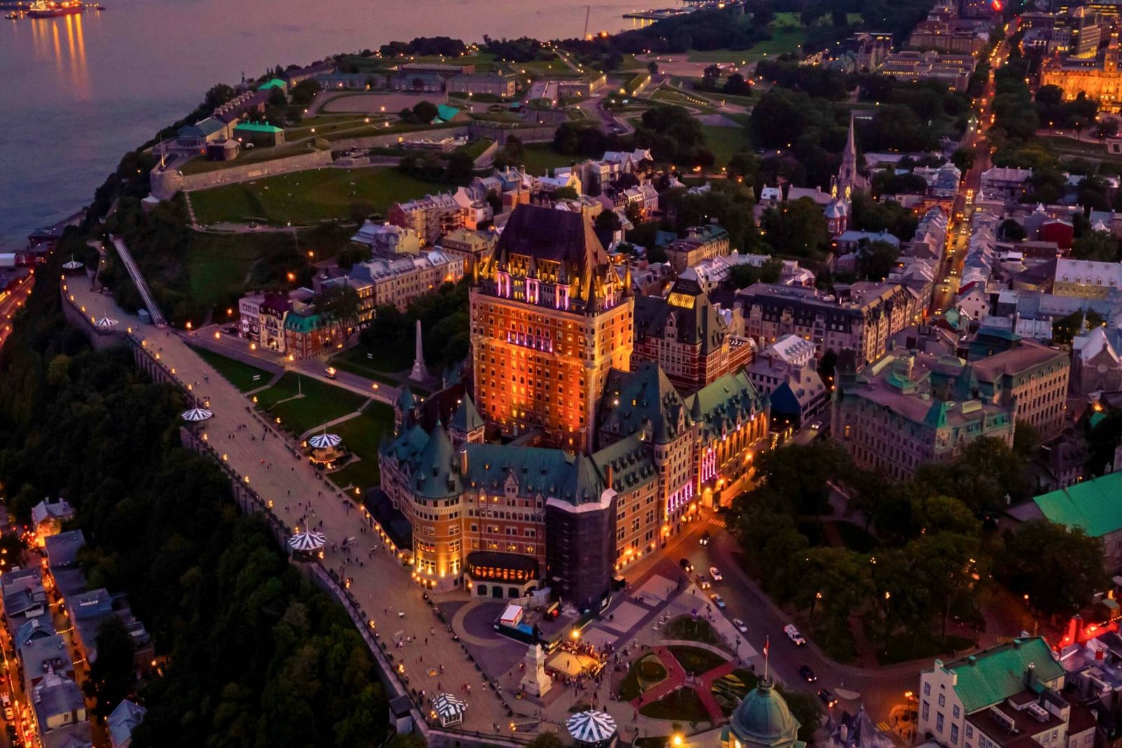 Aerial night view of Fairmont Le Château Frontenac lit up in golden hues, overlooking the Dufferin Terrace and historic Old Québec City by the St. Lawrence River.