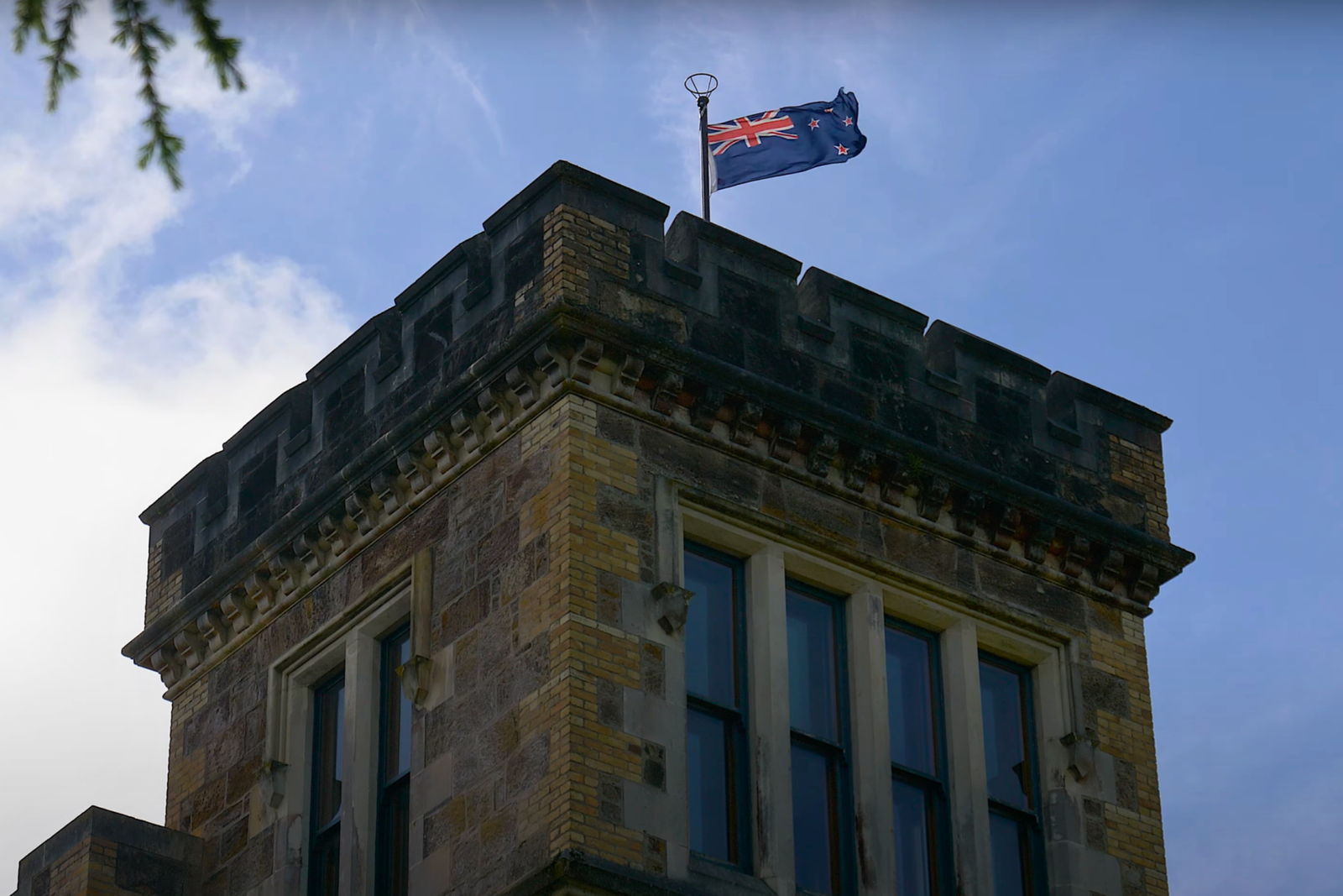 Close-up of the crenellated tower of Larnach Castle with the New Zealand flag waving above, highlighting the castle’s brick and stonework against a blue sky.