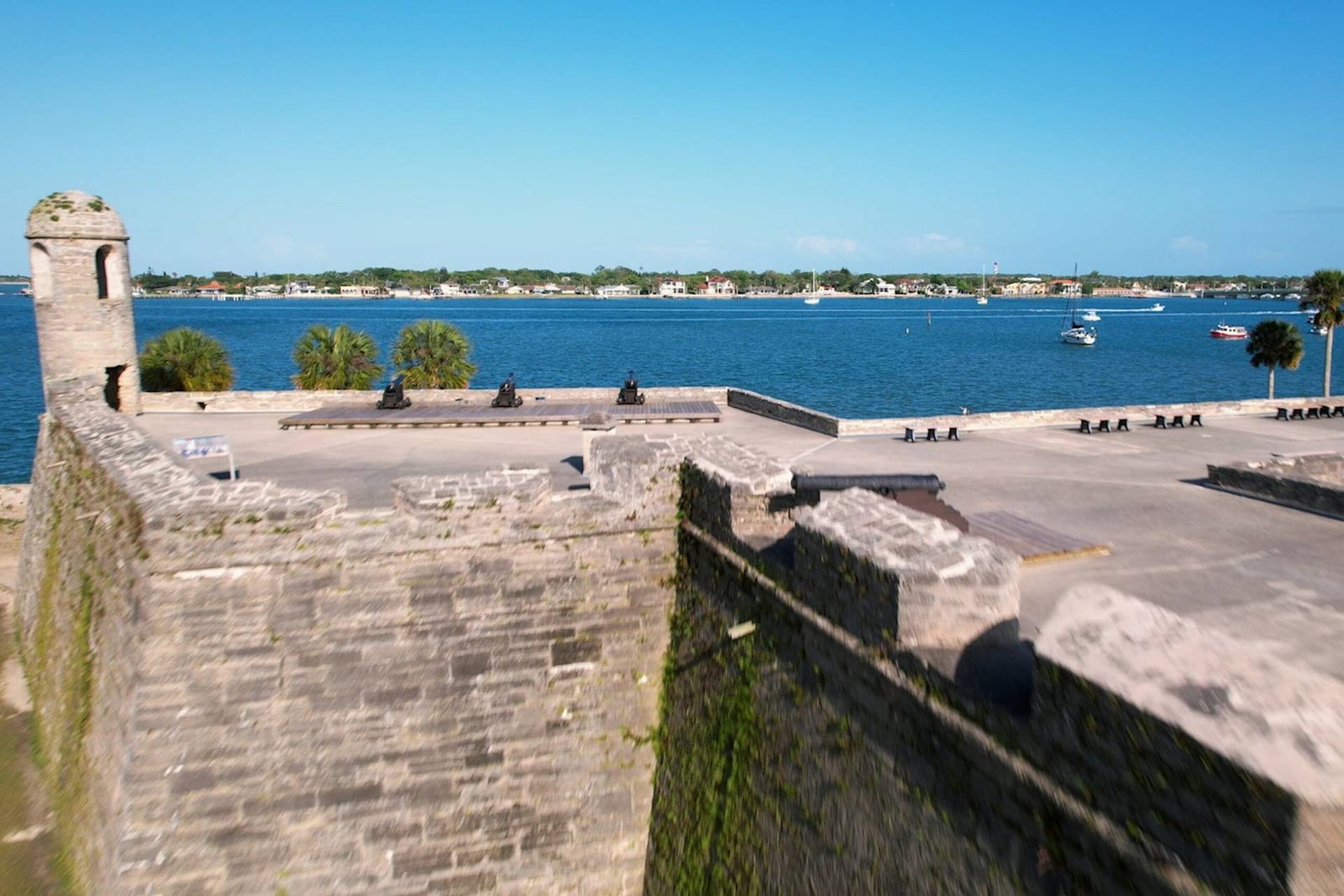 Stone bastions and rooftop cannons of Castillo de San Marcos facing Matanzas Bay in St. Augustine, Florida, with sailboats and palm trees in the background.